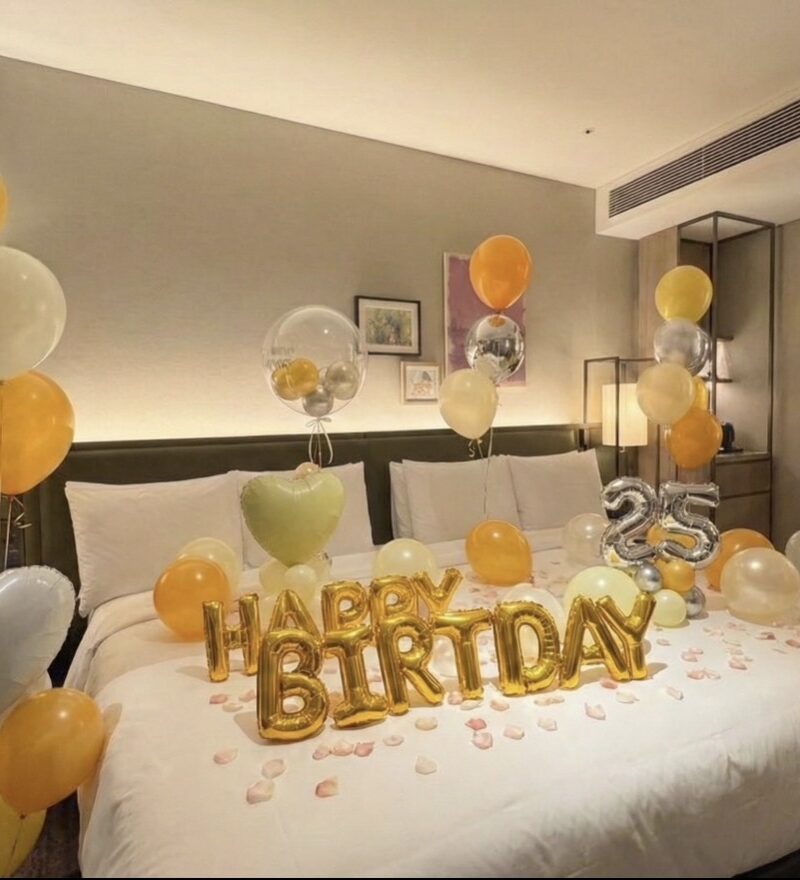 ホテルのベッド全体に施されたバースデーバルーン装飾。ゴールドのHAPPY BIRTHDAYと複数のハート型バルーンがベッドを飾り、周囲に様々な色とサイズのバルーンが配置されている。